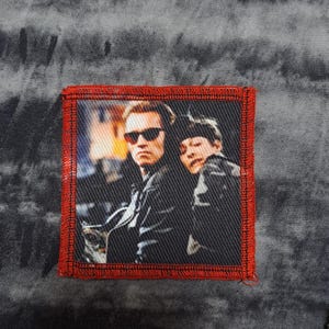 May include: Square patch with a movie still featuring two people, one wearing sunglasses. The patch has a red border and is sewn onto a gray, textured fabric. The image is a close-up of the two people.