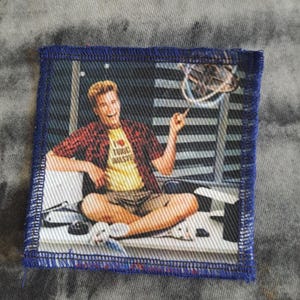 Op de afbeelding: Een vierkante patch met een blauwe rand toont een man in een rood geruit overhemd over een geel shirt met de tekst "I love toxic waste". De man zit met gekruiste benen en wijst omhoog. De achtergrond omvat een ventilator en een gestreepte muur.