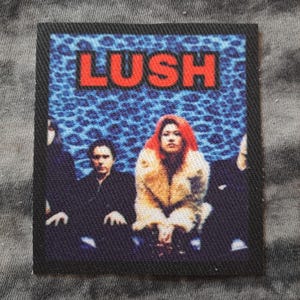 May include: Square patch featuring the band Lush. The word "LUSH" is in red block letters above a photo of the band members. The background is blue with a leopard print design. The patch has a black border.