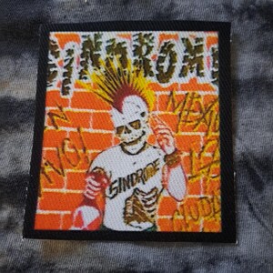 May include: A square patch with a black border featuring a punk rock design. The image shows a skeleton with a mohawk, holding a bottle, in front of a brick wall with graffiti. The text "SINDROME" is on the skeleton's shirt.