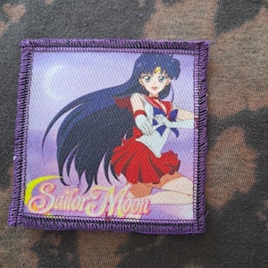 May include: Square patch with a purple border featuring an anime character in a red and white outfit with long dark blue hair. The words "Sailor Moon" are in gold script at the bottom of the patch.