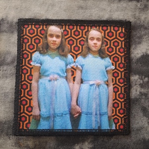 Peut inclure: Un écusson carré représentant deux filles en robes bleues se tenant par la main, sur un motif géométrique orange et noir. L'écusson a une bordure noire, probablement brodée ou imprimée.