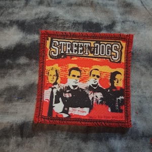 May include: A square, red-bordered patch featuring the text "STREET DOGS" in a stylized font above a graphic of four people. The background is red with a yellow border around the text.