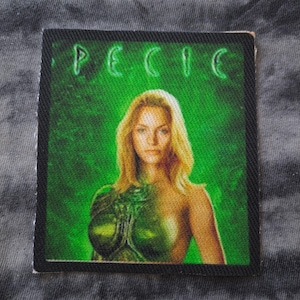 May include: A rectangular patch featuring a woman with blonde hair and a green, alien-like design on her body. The word "SPECIE" is printed in green at the top against a green background. The patch has a black border.