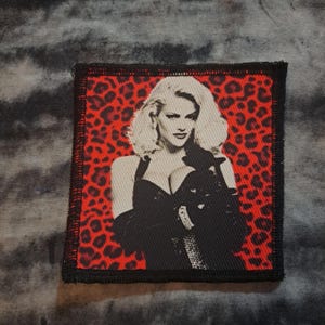 May include: A square patch featuring a black and white image of a woman with blonde hair, set against a red leopard print background. The patch has a black border and is likely for clothing or accessories.