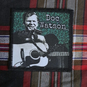 May include: Square patch featuring a black and white image of Doc Watson playing a guitar and harmonica. The patch has a green leopard print background and the text "Doc Watson" in white.