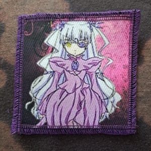 Op de afbeelding: Vierkante patch met een anime-stijl karakter in een paarse jurk, lang wit haar en een paarse ooglap. De patch heeft een paarse gestikte rand en een roze en zwarte achtergrond.