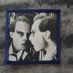 May include: A square patch featuring a black and white image of a man's face, and a profile of the same face. The patch has a dark blue border and is likely an iron-on or sew-on patch.