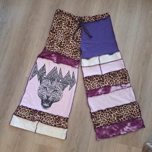 May include: Wide-leg pants featuring a patchwork design of leopard print, solid purple, and light pink fabric. The left leg displays a black and white graphic of a leopard head with the text Def Leppard above it. The pants have a drawstring waist.