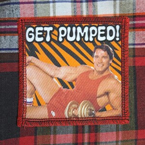 May include: A square patch with the text "GET PUMPED!" above an image of a man in a red tank top holding dumbbells. The background has orange and black stripes. The patch has a red border and is set against a plaid fabric.