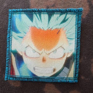May include: Square patch featuring a colorful anime character with spiky blue hair and an orange forehead. The character has an intense expression with visible teeth and determined eyes. The patch has a teal border with a stitched edge.