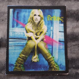 May include: Square fabric patch featuring a seated woman with blonde hair, wearing a yellow outfit. The word "Britney" is in yellow text. The background is blue with a pink streak.
