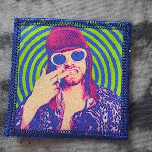 May include: Embroidered patch featuring a portrait of a person with sunglasses, a pink hat, and a leopard print shirt. The background has blue and green concentric circles. The patch has a blue border.