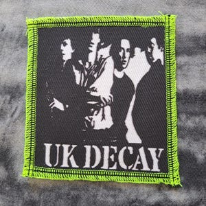 May include: A square patch featuring a black and white image of a band with the text "UK DECAY" in white. The patch has a neon green border with a stitched edge. The patch is on a gray, textured surface.