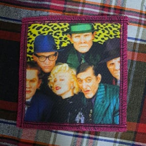May include: A rectangular patch with a magenta border features a group of people with a leopard print background. The people are wearing hats and glasses. One person has blonde hair and red lipstick.