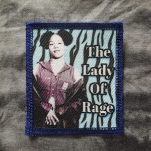 May include: A square patch with a blue border features a portrait of a person with the text "The Lady Of Rage". The background has a blue and white zebra stripe pattern. The person in the portrait is wearing a purple shirt.