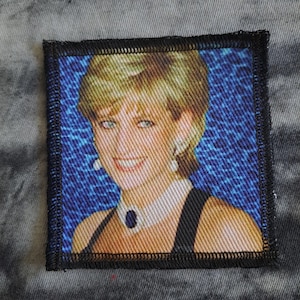 May include: Square fabric patch featuring a portrait of a woman with blonde hair, blue eyes, and a smiling expression. The image is set against a blue patterned background and framed with a black border. The woman is wearing a black dress and a pearl necklace.