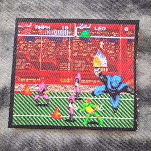 May include: Square fabric patch featuring a pixelated image of a Teenage Mutant Ninja Turtles video game scene. The patch has a black border and depicts characters in action on a green and white striped field, with the names "RAPH" and "LEO" visible.