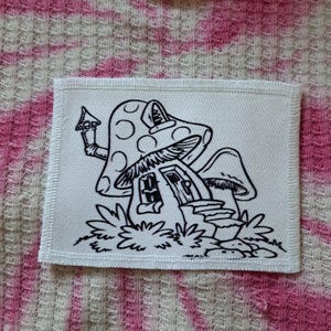 May include: A white fabric patch featuring a whimsical mushroom house illustration in black. The design includes a mushroom-shaped roof with polka dots, a door, windows, and surrounding foliage. The patch has a stitched border.