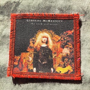 Könnte beinhalten: Quadratischer Aufnäher mit schwarzem Hintergrund und einem Porträt einer Frau mit langen blonden Haaren. Der Aufnäher hat einen rot genähten Rand. Der Text "Loreena McKennitt" ist oben sichtbar. Das Bild hat warme Farben.