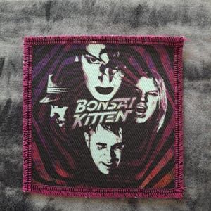 May include: Square patch with a magenta border featuring a graphic of four faces and the text "BONSAI KITTEN" in white. The background has a swirling pattern of purple, black, and red.