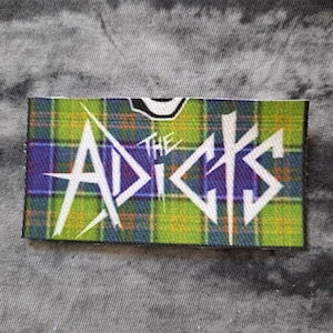 May include: A rectangular patch with a green, blue, and yellow plaid pattern. The text "THE ADICTS" is in white, stylized lettering. The patch is likely for clothing or accessories.