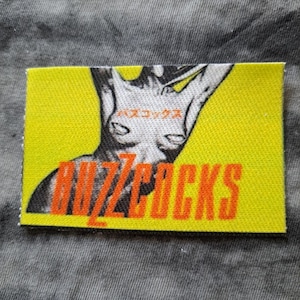 May include: A rectangular yellow patch with the word "BUZZCOCKS" in orange block letters. A black and white illustration of a figure is above the text. Japanese text is above the figure.