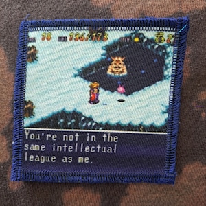 May include: Square patch with a blue stitched border featuring a pixelated video game scene. The scene depicts a character in a red and gold outfit, with text that reads, "You're not in the same intellectual league as me."