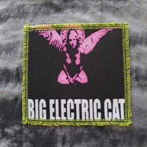 May include: Square patch with a black background and a pink graphic of a winged figure. The patch has a neon green border and the words "BIG ELECTRIC CAT" in white block letters.