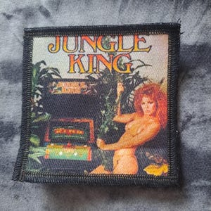 Peut inclure: Écusson carré avec les mots "JUNGLE KING" en lettres dorées. L'écusson représente une femme aux cheveux roux dans un cadre de jungle. L'écusson a une bordure noire.