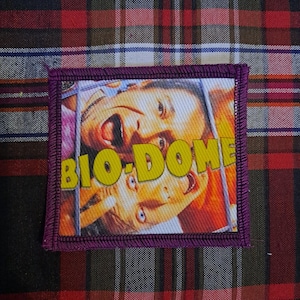 May include: Square patch with a purple border featuring a movie still with the text "BIO-DOME" in yellow. The image shows two people with surprised expressions. The patch is on a plaid fabric background.