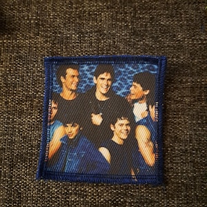 May include: A square fabric patch featuring a printed image of five young people with dark hair, posed together against a blue textured background. The patch has a dark blue stitched border and a visible woven fabric texture throughout the design.