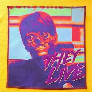 Puede incluir: Parche cuadrado con una imagen colorida de una cara con forma de calavera y el texto "THEY LIVE" en morado. El parche tiene un borde rojo y está sobre un fondo amarillo.