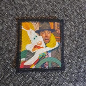 Pode incluir: Um emblema bordado retangular com um gráfico colorido de um coelho de desenho animado branco com orelhas grandes e uma expressão de surpresa ao lado de um personagem humano usando um chapéu fedora escuro, com uma borda preta costurada.
