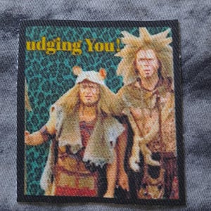 Könnte beinhalten: Rechteckiger Aufnäher mit einem farbenfrohen Bild von zwei Personen in Kostümen. Der Text "Judging You!" ist gelb gedruckt. Der Hintergrund ist ein teal-grünes Muster.