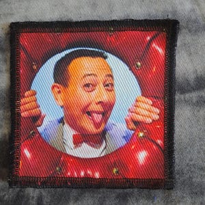 May include: Square fabric patch featuring a portrait of a man with his tongue sticking out, framed by a red background. The man is wearing a bow tie and a light blue shirt. The patch has a black border.