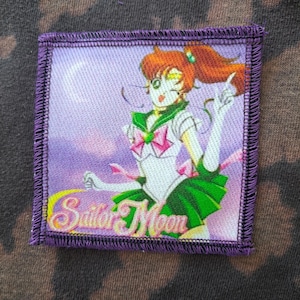 Puede incluir: Un parche cuadrado con borde morado que representa un personaje de Sailor Moon con un atuendo verde y blanco con un lazo rosa. El parche tiene las palabras "Sailor Moon" en amarillo y verde.