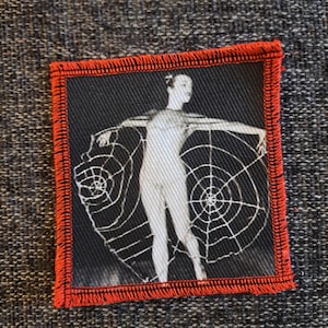 May include: A square fabric patch featuring a black and white photographic print of a person standing with arms outstretched, holding large white spider web patterns. The patch has a thick, bright red stitched border around the entire edge.