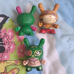 May include: Three colorful vinyl bunny figurines. One is green with pink accents, resembling a cactus. Another is tan with a monkey face and floral details. The third is green with a luchador mask.