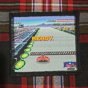 May include: A square patch featuring a retro video game racing scene with the word "READY" in yellow. The patch has a black border and is set against a red and black plaid background.