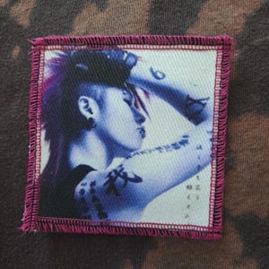 May include: Square patch with a portrait of a person with purple hair and tattoos, in shades of blue and white. The patch has a pink stitched border and is set against a mottled brown background.