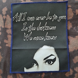May include: A rectangular patch with a black background and a blue border. The patch features the text "All I can ever be to you Is the darkness We once knew" in white script. Below the text is a black and white image of a woman's eyes.