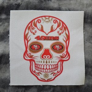 Puede incluir: Un parche cuadrado blanco con un diseño de calavera de azúcar rojo y dorado. La calavera tiene las palabras "49ERS" en la frente y el logotipo del equipo en las cuencas de los ojos. Sobre un fondo gris y blanco.