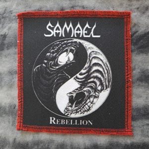 May include: Square black patch with a red stitched border. The patch features a yin-yang design with snake-like creatures and the word "SAMAEL" above the design. The word "REBELLION" is below the design.