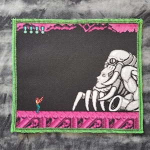 May include: Square patch featuring a retro video game scene with a pixelated character shooting at a large, white, alien-like creature. The patch has a black background with pink and green accents.