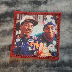 May include: A square patch with a red border showing a colour image of two people. One person wears a baseball cap and a jacket with patches. The other person wears a blue and white hat and a green shirt. The background is a tie-dye pattern.