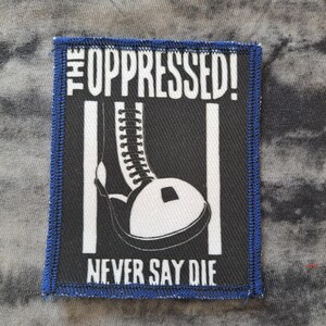 May include: A black and white patch with a blue border. The patch features the text "THE OPPRESSED!" above an illustration of a boot and the text "NEVER SAY DIE".