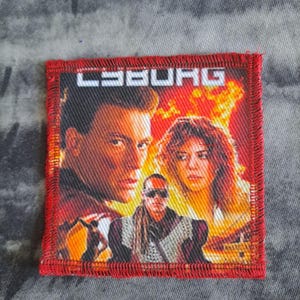May include: Square patch with a red border featuring a movie poster design. The poster has a fiery background and the word "CYBORG" in white block letters. The image depicts three people, with two in the foreground.