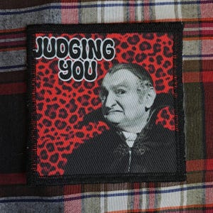 May include: Square patch with the text "JUDGING YOU" in white letters with a black outline. The background is red with black leopard spots. A black and white image of a man is in the lower right corner. The patch has a black border.