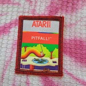 May include: A rectangular Atari Pitfall! patch with a red border. The patch features the Atari 2600 logo, the game title "Pitfall!", and a colorful graphic of the game's environment, including a rainbow, trees, and a pond with crocodiles.